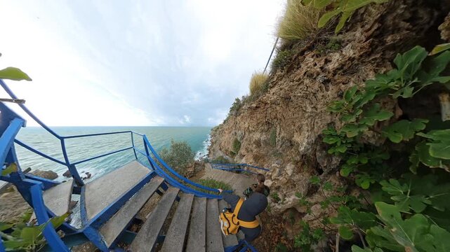 A fisheye view of a man and a woman descending a blue staircase winding along a steep cliff, surrounded by rugged rocks and greenery, with the sea visible ahead