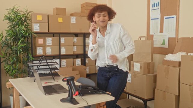 Woman dancing with arms raised beside stacked cardboard boxes and a packing desk with laptop and scanner in an office building; business success joy.