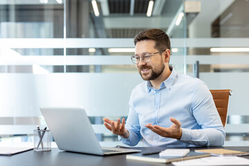 Fototapeta premium Young businessman wearing glasses and a blue shirt, smiling and gesturing during a virtual conference or remote work video call on a laptop from his modern office desk