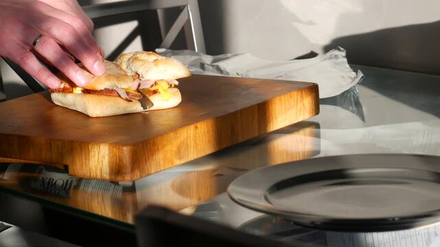 Man cutting sandwich on wooden cutting board with knife. Kitchen scene with fresh food preparation for meal. Culinary process of slicing bread with meat inside.