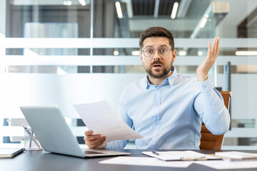 Obraz premium Stressed male employee holding a document, looking confused and frustrated with an open mouth gesture, sitting at his office desk with a laptop and papers