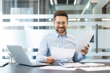 Obraz premium Smiling businessman reviewing business documents, financial reports, and contracts while working at a desk with a laptop in a modern office, planning strategies for success