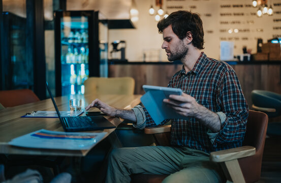 A man works on a laptop and tablet in a cafe, reviewing documents and data for business. He sits at a table in a cozy coffee shop using multiple devices for remote work.