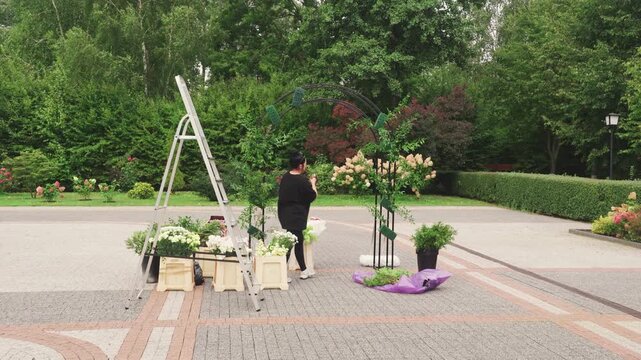 Laddermounted florist arranges leafy backdrop. Potted blooms and crates decorate garden arch with blooms. Florist installs arch outdoors by adjusting frame amidst lush green foliage and crates