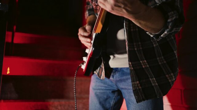Close-up of Electric Guitar Player with Dramatic Red Lighting. Musician hands playing a black and white electric guitar. Atmosphere with vibrant red backlight, featuring an amplifier in the background