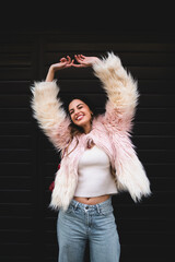 Joyful young woman raising arms and laughing against dark background, happy girl in stylish pink faux fur coat expressing freedom and success, emotional portrait of real people, copy space.