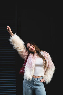Joyful woman raising one arm in celebration against black background, happy girl in fluffy pink coat expressing freedom and success, emotional connectivity and urban lifestyle, copy space.