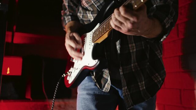 Close-up of Electric Guitar Player with Dramatic Red Lighting. Musician hands playing a black and white electric guitar. Atmosphere with vibrant red backlight, featuring an amplifier in the background