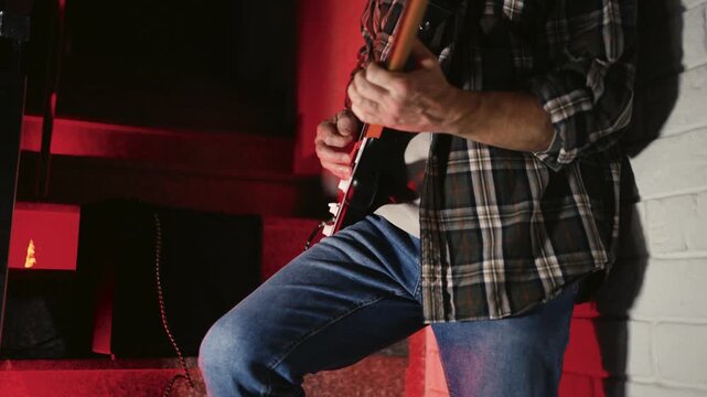 Close-up of Electric Guitar Player with Dramatic Red Lighting. Musician hands playing a black and white electric guitar. Atmosphere with vibrant red backlight, featuring an amplifier in the background