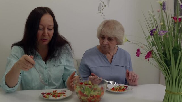 Two white women eating salad at table, daughter caregiver and elderly mother share quiet, slightly tense mealtime, colorful glass bowl of salad, forks and plates, floral vase on side,