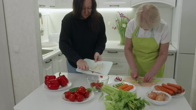 Cooking team with fresh ingredients. Two women chop vegetables demonstrating teamwork. Two women of Caucasian ethnicity collaboratively slicing vegetables on vibrant kitchen counter