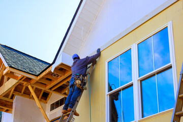 Worker is using ladder to place siding on new building at construction site.