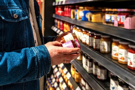 White man reading jar label on shelf, denim sleeve and hands visible, close shelf lighting, checking ingredients and nutrition for dietary choice, careful comparison before purchase