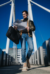 Young man reads a map while walking across a modern pedestrian bridge carrying a black briefcase....