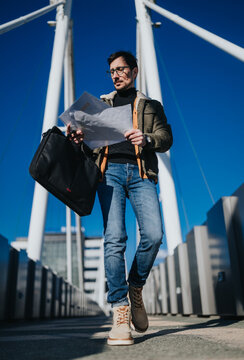 Young man reads a map while walking across a modern pedestrian bridge carrying a black briefcase. Casual urban commuter navigates with paper directions against a clear blue sky.