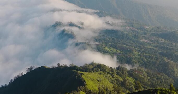 Aerial view of the vibrant green mountain range, partially concealed by a blanket of soft, white clouds, creating a serene and ethereal landscape, Bali, Indonesia.