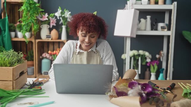 Woman florist smiles and raises arms at her laptop amid flowers in a busy building; online sales joy.