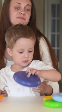 A woman observes a boy as he plays with colorful toys on a table. The boy interacts with a ring stacker toy while the woman watches with interest.