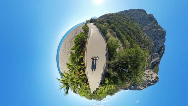 A tiny planet aerial view showcasing a curved pathway bordered by lush greenery and cliffs, with two people walking near the beach under a vibrant blue sky