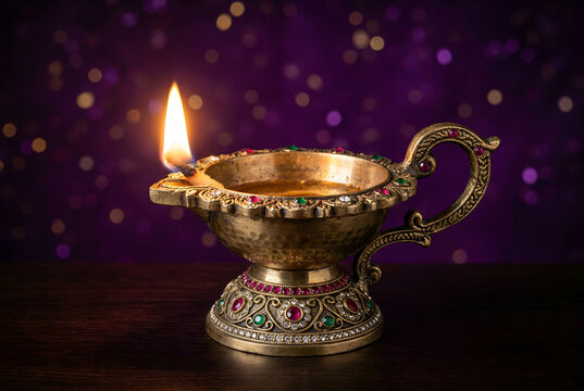 Decorative brass oil lamp diya with handle, lit flame and colorful jewels against blurred purple background symbolizing light