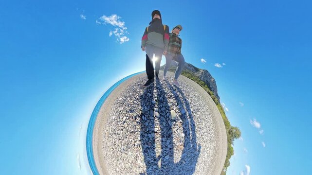 A tiny planet effect showing a couple walking together on a pebbled beach, casting long shadows under a bright sun with turquoise water and cliffs in the background