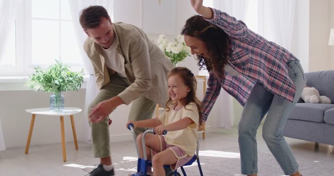 Happy parents and daughter play indoors together. Mother and father having fun with child girl guide her on a tricycle in living room. Parents build confidence and safety. Warm family time at home.