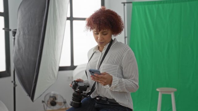 Woman photographer holding camera and smartphone to ear in studio with green screen, softbox and stool; focused.
