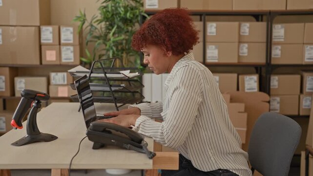 Woman typing on laptop with hand on lower back at building desk amid stacked cardboard boxes, barcode scanner and paperwork; work stress.