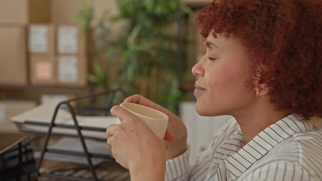 Woman cupping mug to nose with closed eyes, hands holding coffee while smiling among stacked cardboard boxes and parcel shelves in an office building; contentment.
