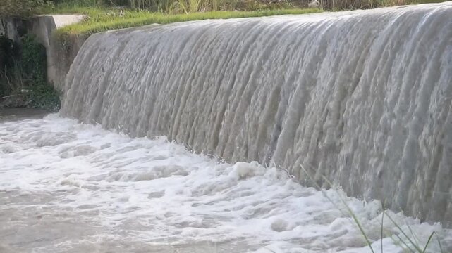 Muddy river water flowing over a small dam, creating a textured cascade with splashing foam and dynamic motion, ideal for flood, environment, and water management concepts.
