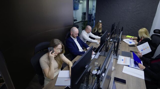 A team of business professionals works at their desks in a contemporary office setting. Colleagues collaborate on projects, utilizing computers and analyzing various documents and data.