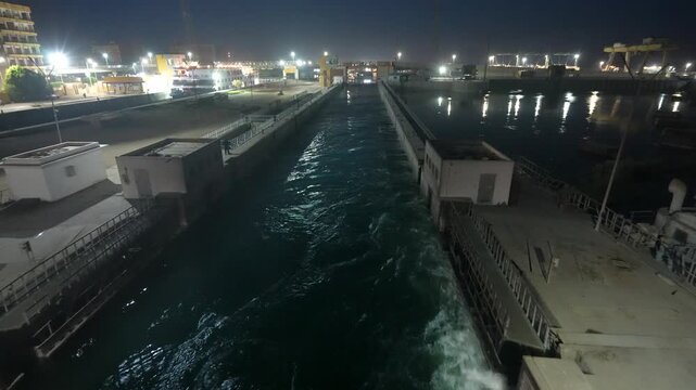 A night view of the Esna Lock in Luxor, Egypt, showing a ship navigating through the water channel with illuminated industrial structures.
