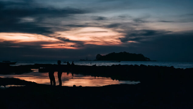 Silhouettes of people on a rocky coast during a fiery sunset.