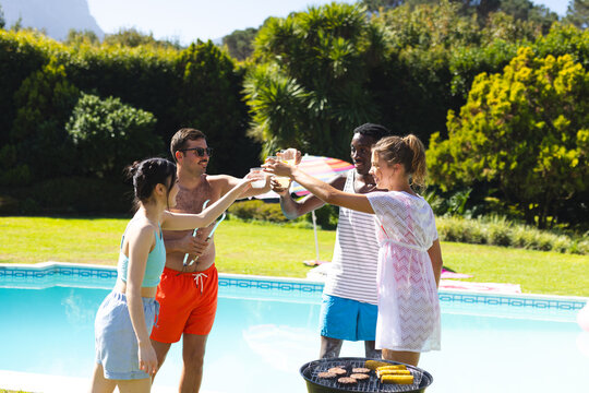 Toasting diverse friends raising glasses beside backyard pool, with barbecue grill and umbrella
