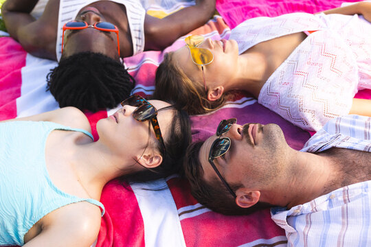 Reclining diverse friends soaking up sun on colorful beach towels on grassy lawn, with sunglasses