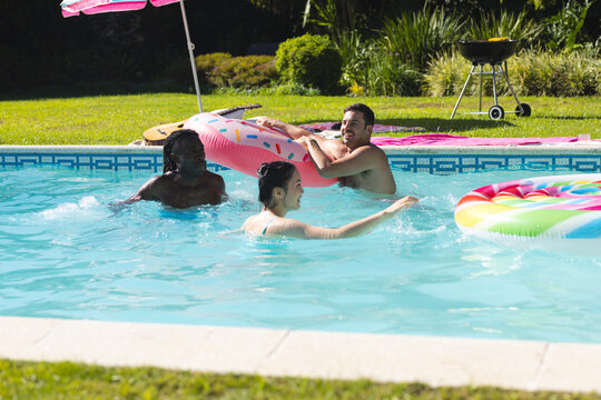 Playing Diverse friends splashing in backyard pool, with pink donut float and guitar