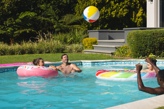 Laughing Diverse friends splashing in backyard pool, with pink donut float and rainbow tube