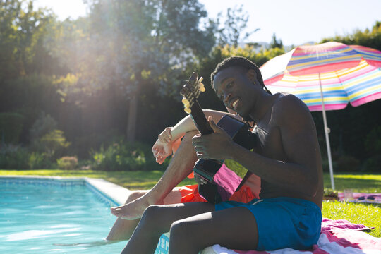 Sitting diverse male friends strumming acoustic guitar poolside under striped umbrella, copy space