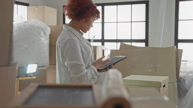 Woman holding tablet and tapping screen while checking boxes in a building filled with moving supplies; planning determination.