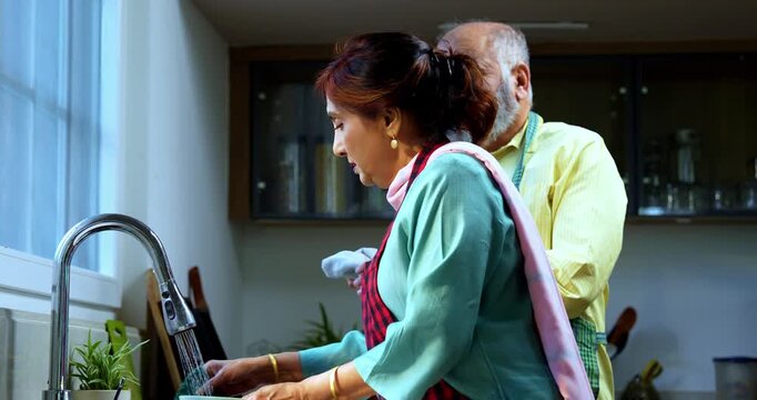 Retired senior Indian Asian old age couple together washing dishes at a modern kitchen platform; showcasing teamwork, daily household chores, and togetherness in a contemporary home setting.