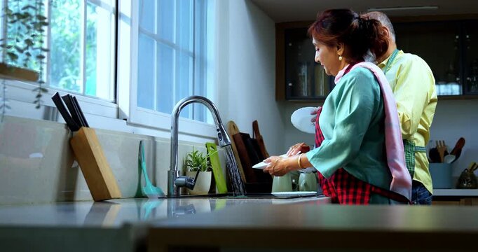 Retired senior Indian Asian old age couple together washing dishes at a modern kitchen platform; showcasing teamwork, daily household chores, and togetherness in a contemporary home setting.