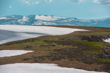Scenic minimalist view from snowy field to sunlit snow cornice against large snow-capped mountain range silhouette. Alpine minimal landscape with snowfield on top in sunlight. Thaw in high mountains. © Daniil