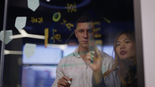 Diverse business professionals are actively engaged in brainstorming and strategizing on a transparent glass board.