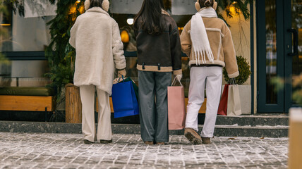 Fototapeta premium Three friends enjoying winter shopping, carrying colorful bags outside a store