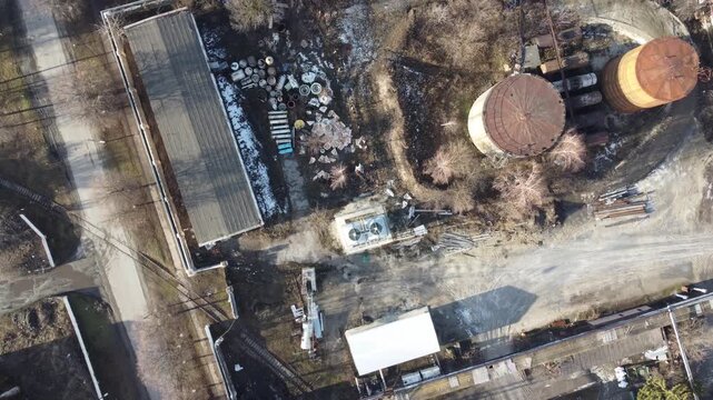 An aerial top-down view reveals an old industrial plant with multiple rusty storage tanks and dilapidated buildings. The scene conveys a sense of abandonment and disuse.