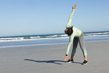 Fototapeta premium Mid adult Asian woman doing upright side-bend on beach with waves, tracker and sneakers, copy space