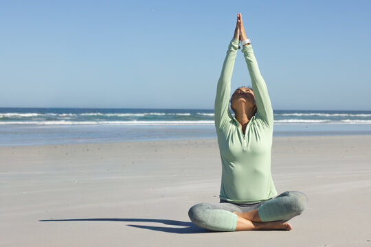 Mid-adult Asian woman doing yoga, arms raised cross-legged on sand in athletic top, leggings, watch