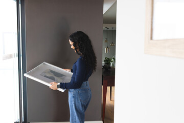 Fototapeta premium Framed artwork is leaning near tall glass door against dark gray wall, wooden table visible