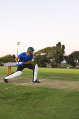 Obraz premium Adult male cricket batter holding wooden bat in helmet on pitch at dusk with stumps behind