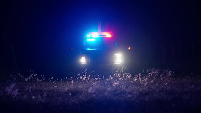 Police vehicle with bright headlights and flashing red and blue lights in night darkness creating moody cinematic emergency scene with strong glow and atmosphere
